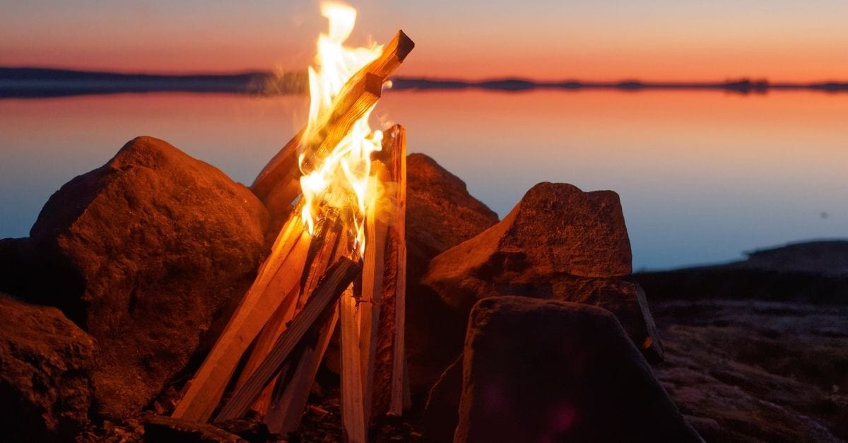 Ein Lagerfeuer an einem See während eines roten Sonnenuntergangs.