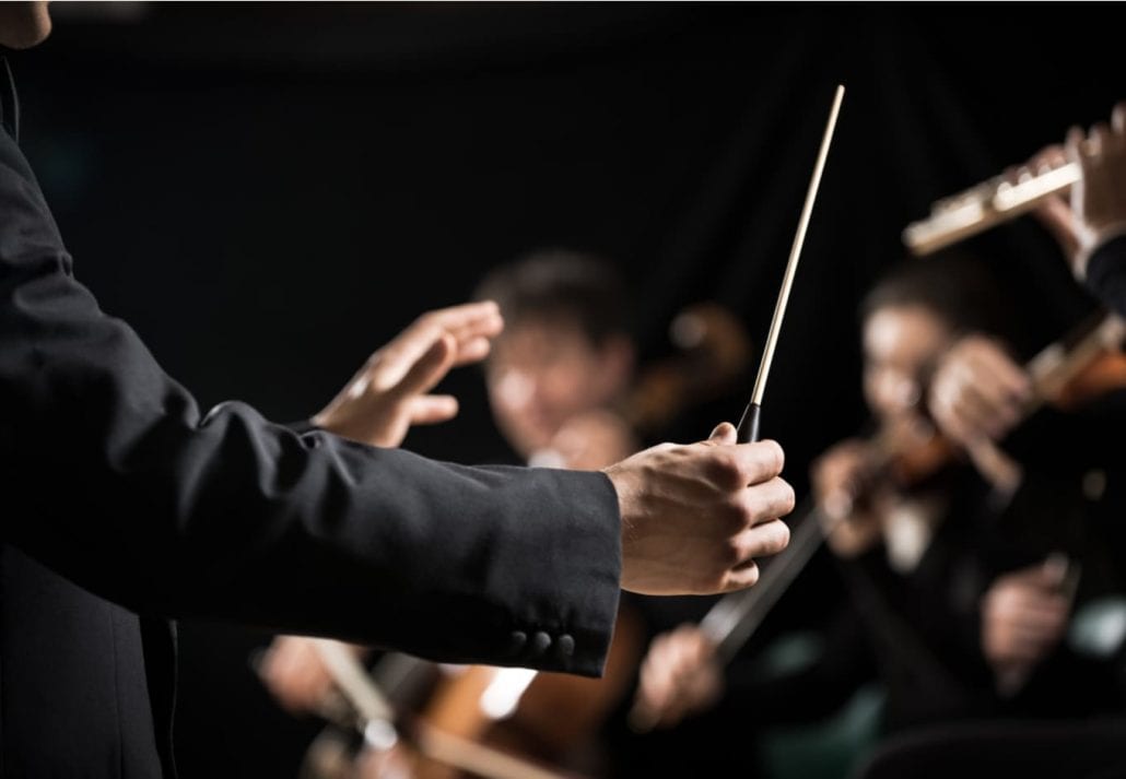 Maestro conducting symphony orchestra with performers in the background, close-up of hands.