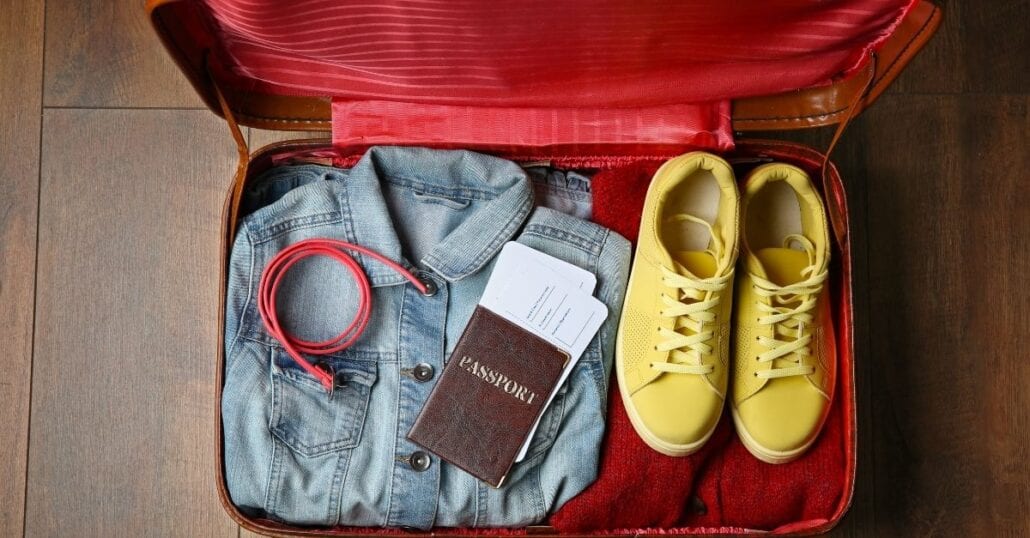 A jacket, an USB cable, a passport, flight tickets and a pair of sneackers inside a red travel suitcase.