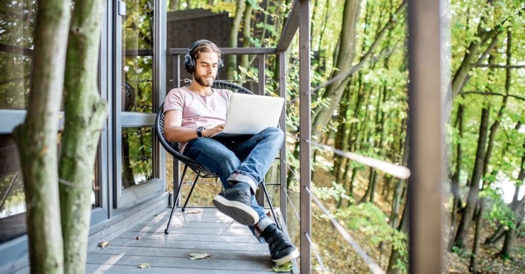 A youn man working on his laptop on a cabin wood deck.