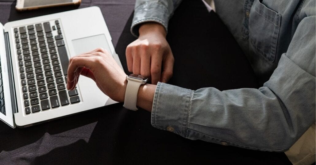 Remote employee working on his laptop and checking the time on his watch. 