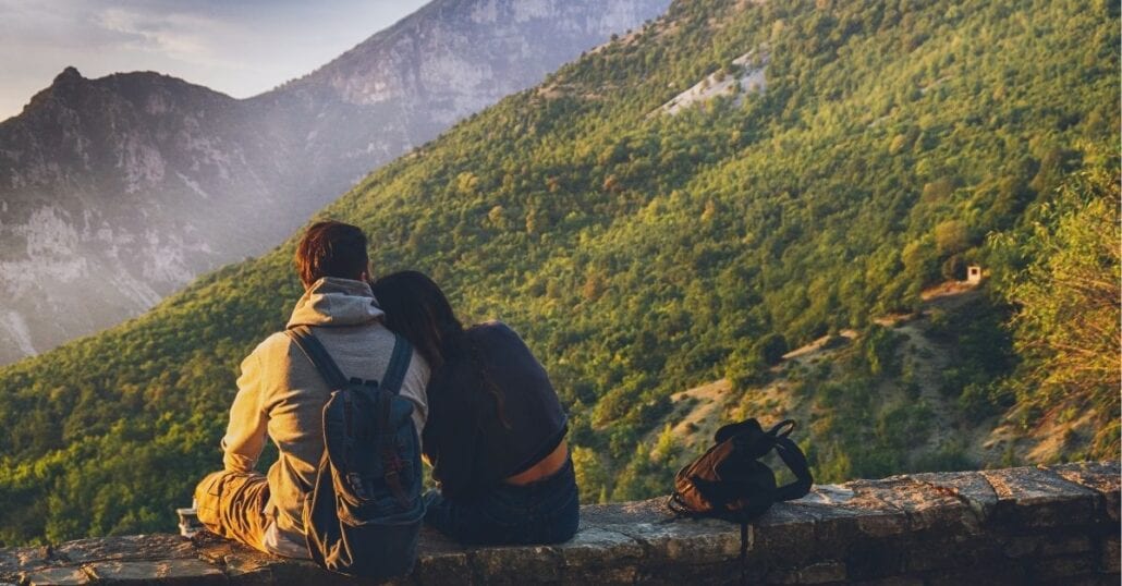 Adventurous-couple sitting and contemplating a a gigantic verdant mountain.