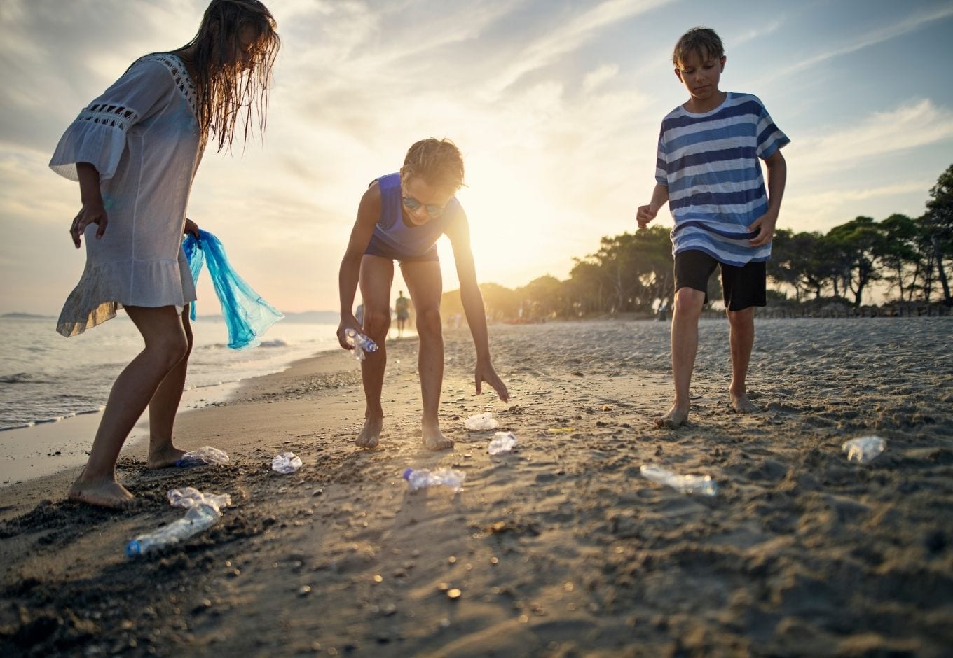 Three kids cleaning up a beach during the sunset.