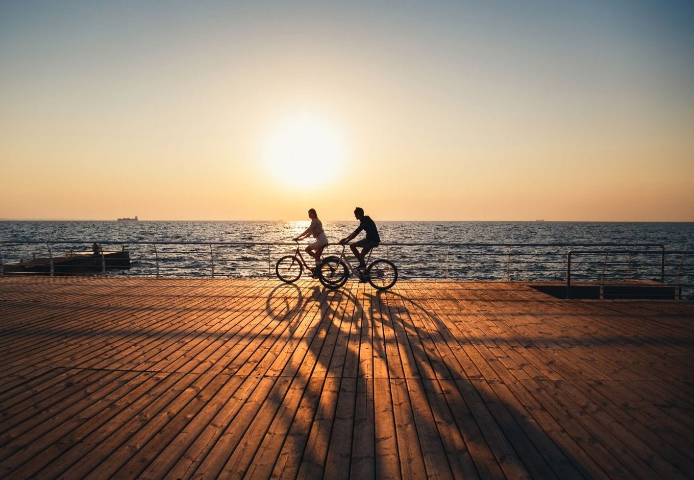 A woman and a man cycling over a wooden deck by the ocean at sunset.