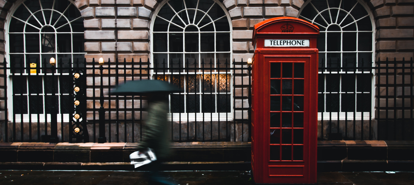 Man holding an umbrella and walking by a red telephone booth on a rainy and cloudy day in London, UK.