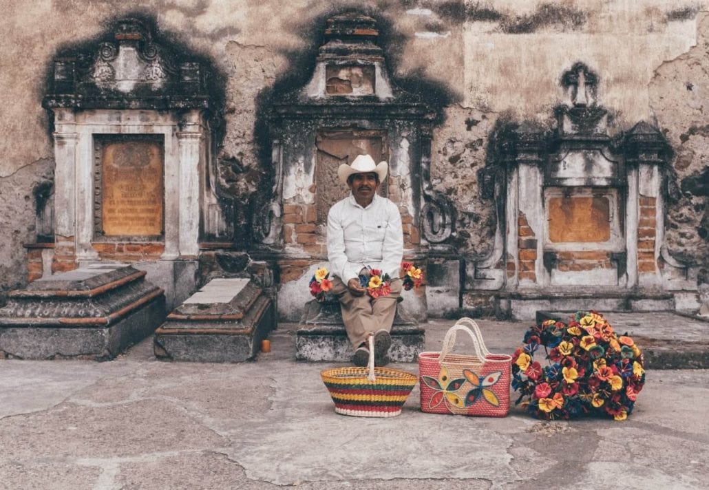 Portrait of a Mexican man peacefully sitted on a grave, in Mexico, and holding colorful flowers.
