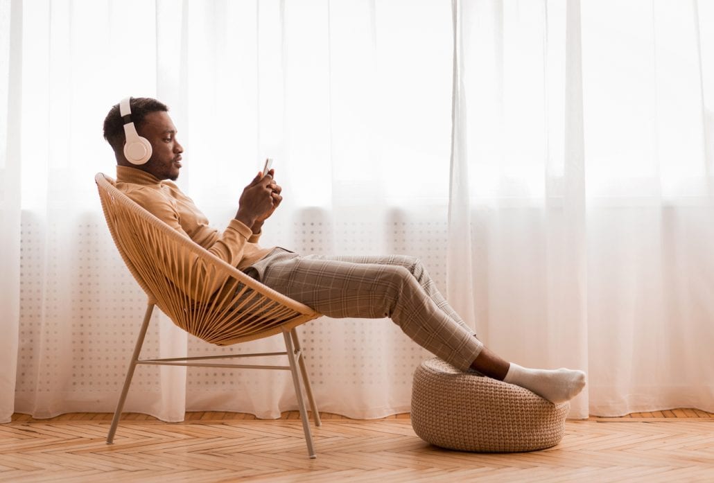 man-with-headphones-on-travel-podcasts | CuddlyNest Travel Blog Relaxed Man Listening To Podcast Sitting On Modern Chair Against Window Indoor