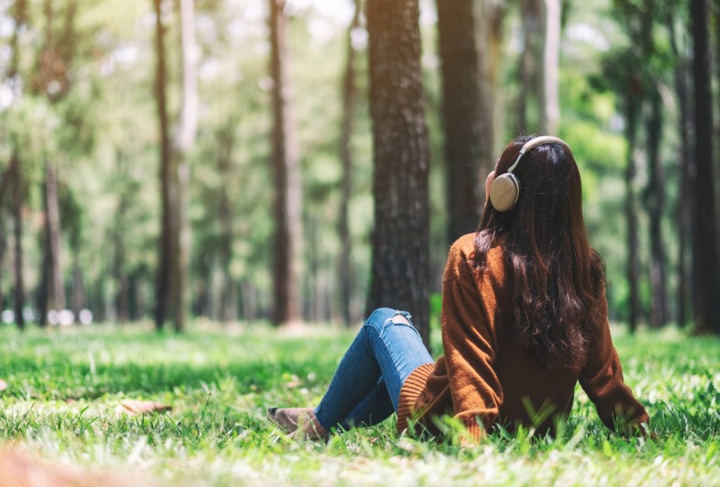 woman-in-headphones-travel-podcasts | CuddlyNest Travel Blog Young woman listening to music with headphones on while sitting at the grass at a park.