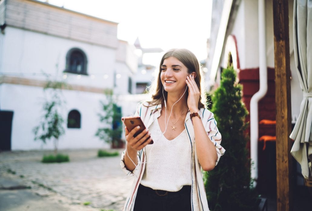 woman-listening-to-travel-podcast | CuddlyNest Travel Blog Smiling girl in earphones listening to music on her smartphone.