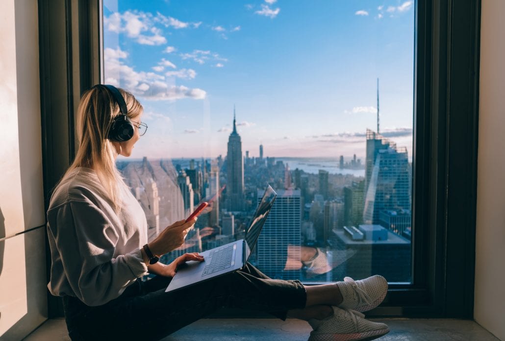 woman-with-headphone-travel-podcasts | CuddlyNest Travel Blog Blond woman with headphones on, a laptop on her lap, sitting on the windowsill and looking at New York's skyline.