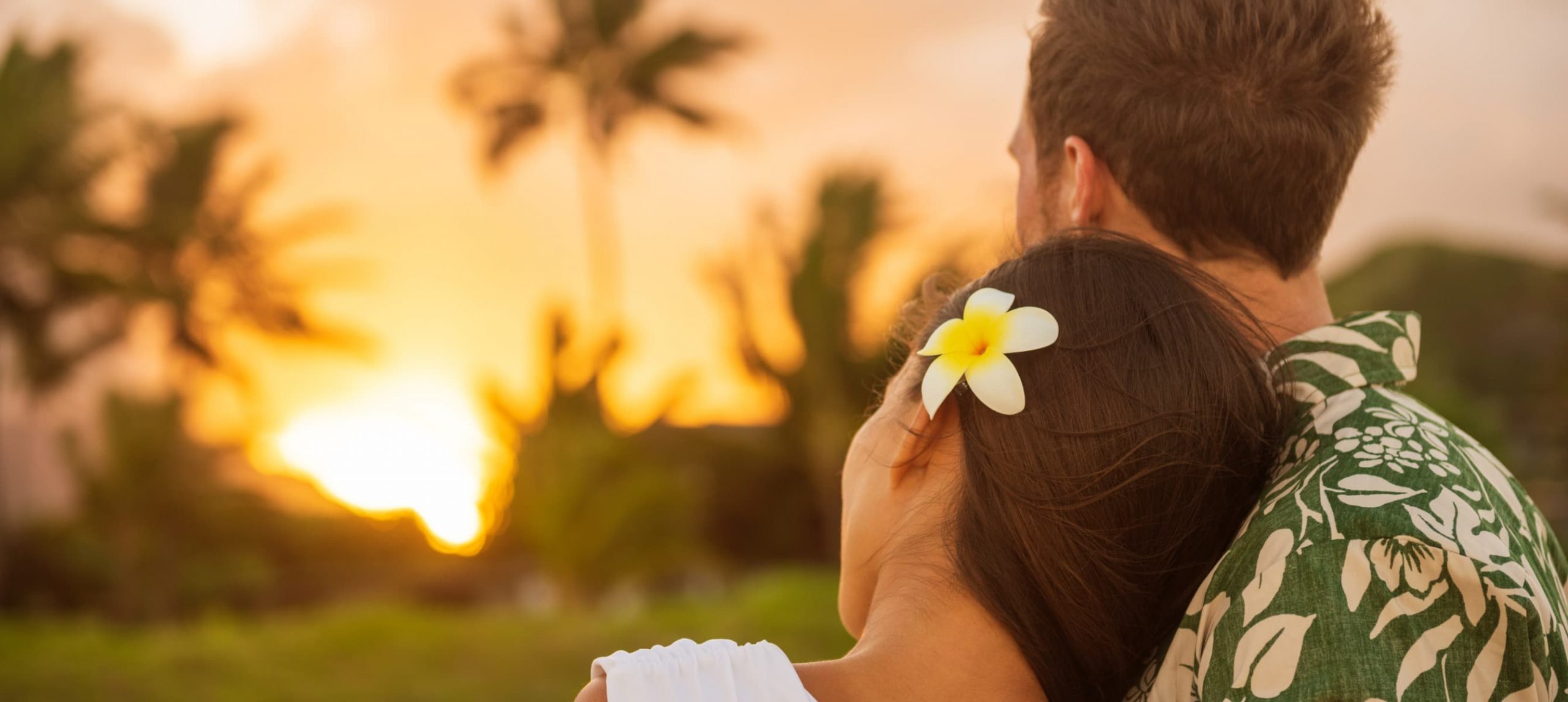 Romantic couple relaxing watching sunset on beach stroll view from back. Woman resting head on lover's shoulder on honeymoon vacation travel in summer Hawaii destination. Newlyweds people.