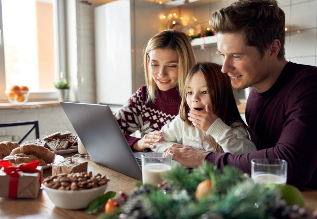 Happy young parents with cute excited small kid daughter using laptop computer at home sitting at Christmas table