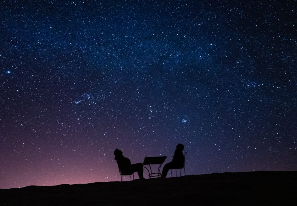 young couple sitting at a table on a desert dune while talking, relaxing and observing the stars and the milky way above them