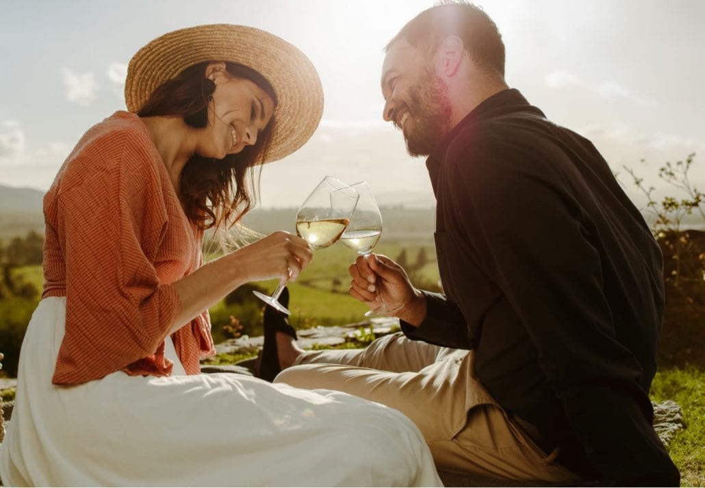 Couple sitting on grass in a vineyard toasting wine. Smiling woman in hat sitting with her boyfriend drinking wine and talking to him.