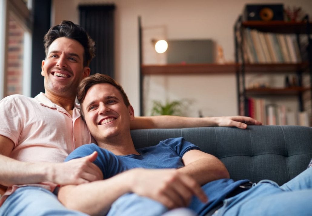  Male Couple Lying On Sofa At Home Watching TV And Relaxing Together