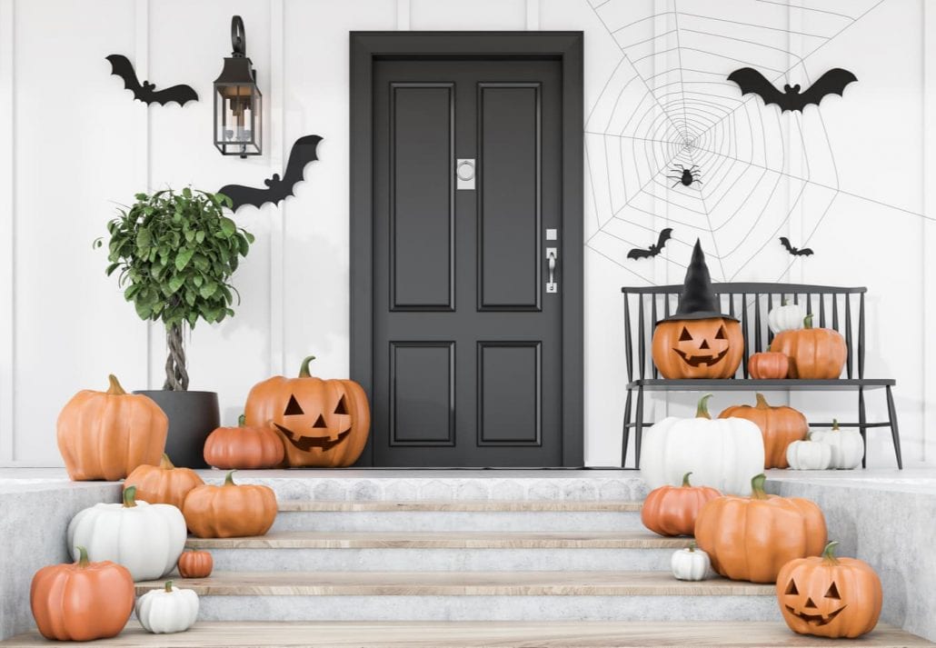Carved pumpkins, bats and spiders on stairs and bench near modern house with black front door, tree in pot and white walls. Concept of halloween.
