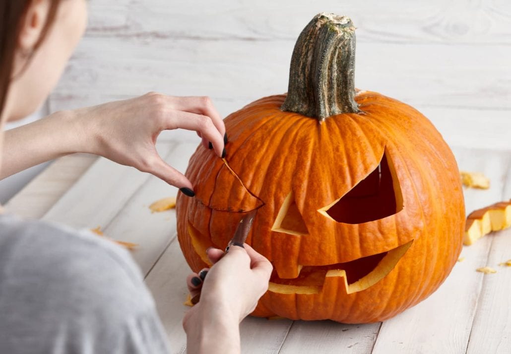 Woman carving big orange pumpkin into jack-o-lantern for Halloween holiday decoration on white wooden planks, close up view