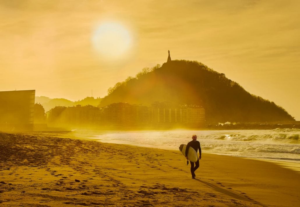 A surfer walking on the Zurriola Beach at sunset with the Monte Urgull in the background. San Sebastian, Basque Country, Guipuzcoa.