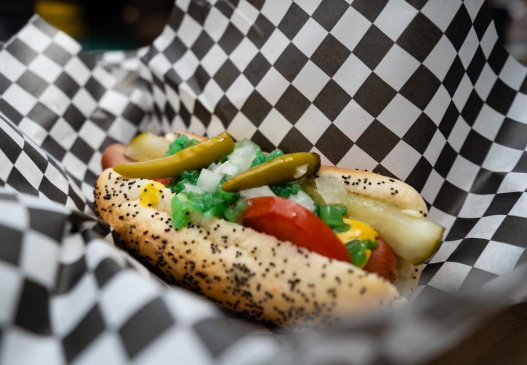 Chicago Style Hot Dog in checkered wrapper served with sport pepper, pickle, tomato, onion, sweet relish, yellow mustard, all-beef frankfurter, on a poppy seed bun.