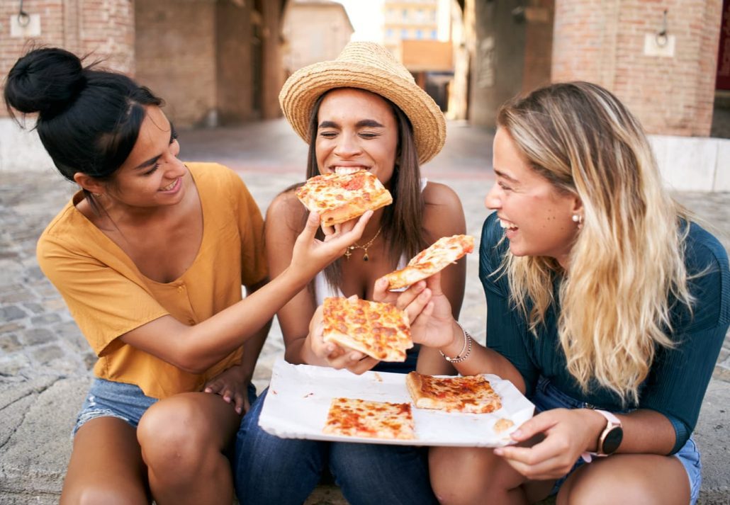 Three young women laughing while eating pizza together in Italy.