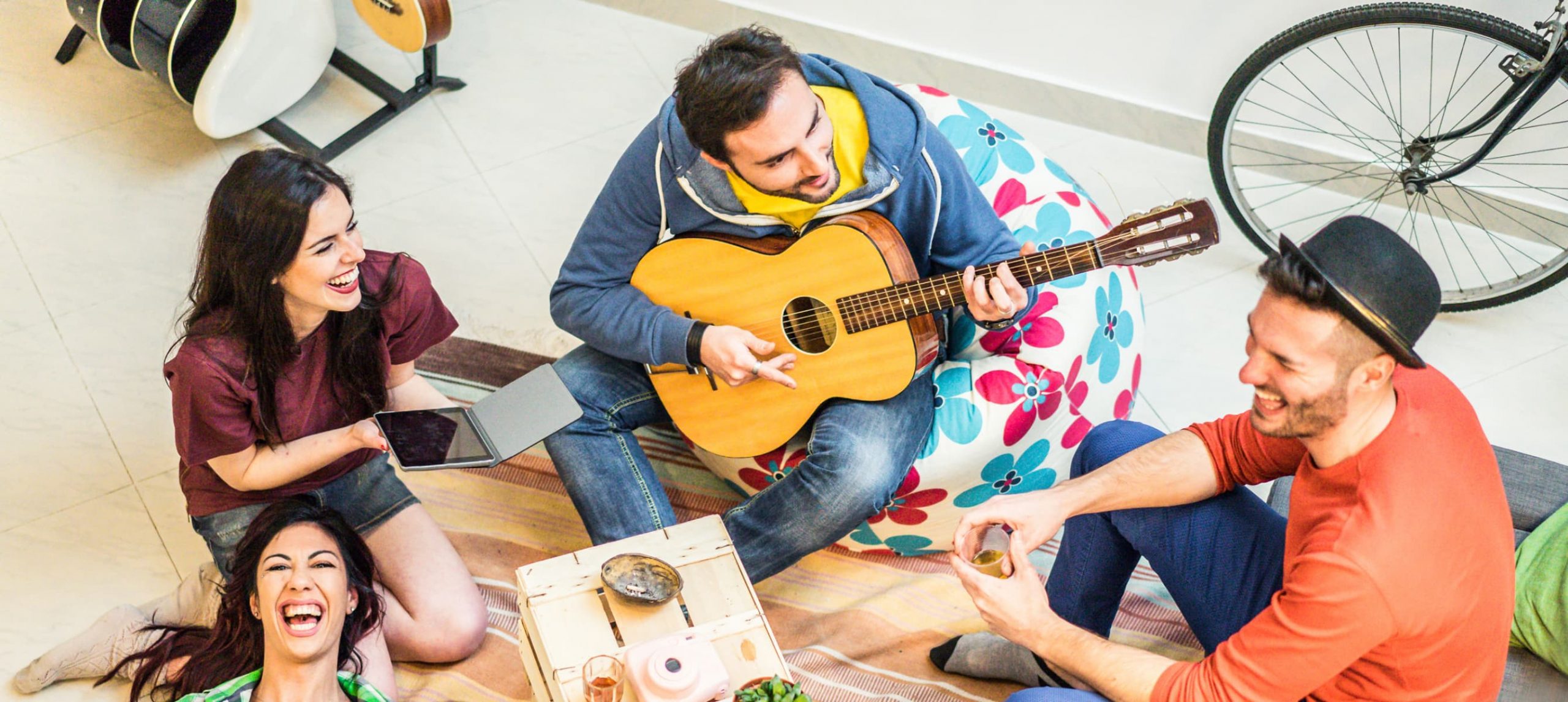 A group of friends laughing while playing the guitar and singing while at a hostel.