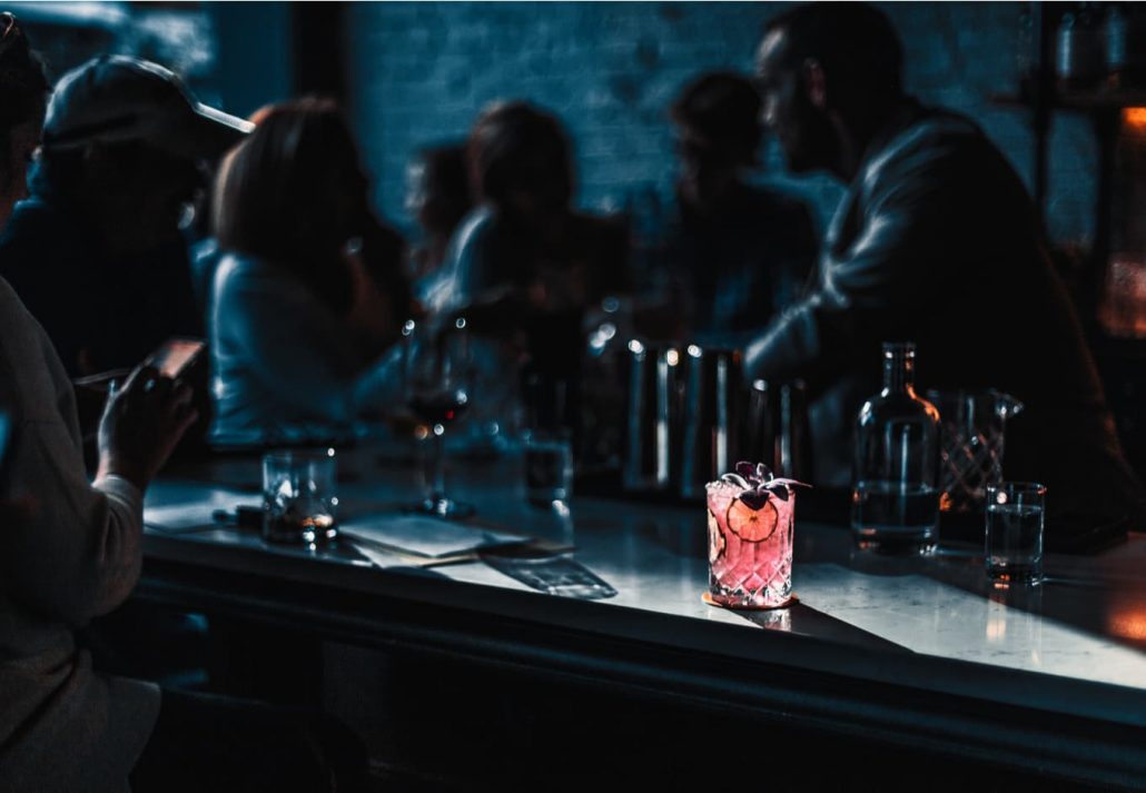 berlin-gay-party-scene-zum-schmutzigen-hobby | CuddlyNest Travel Blog People chatting at the table in a dark bar with a pink cocktail in front of them
