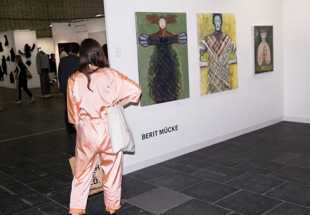 A female posing with her back turned to camera, standing in front of artworks