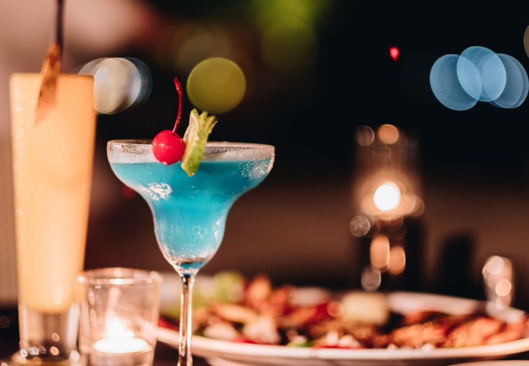 A blue cocktail on a table with food served behind it
