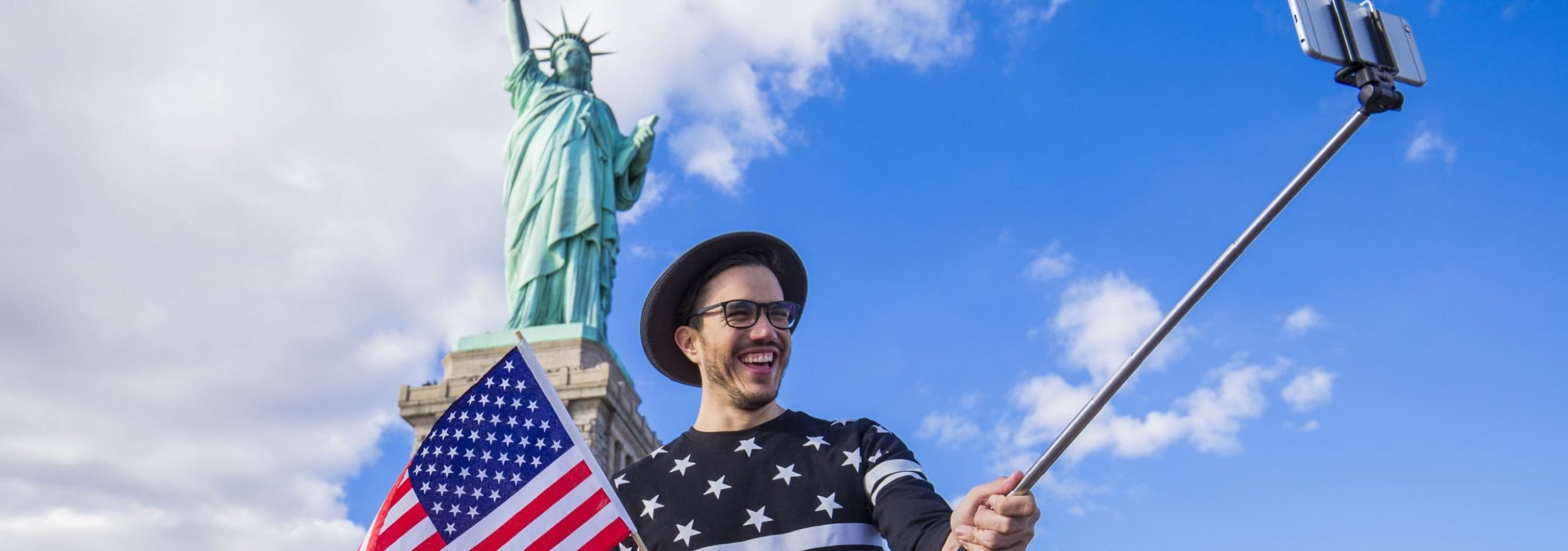 a tourist taking a selfie with a USA flag in front of the Statue of Liberty