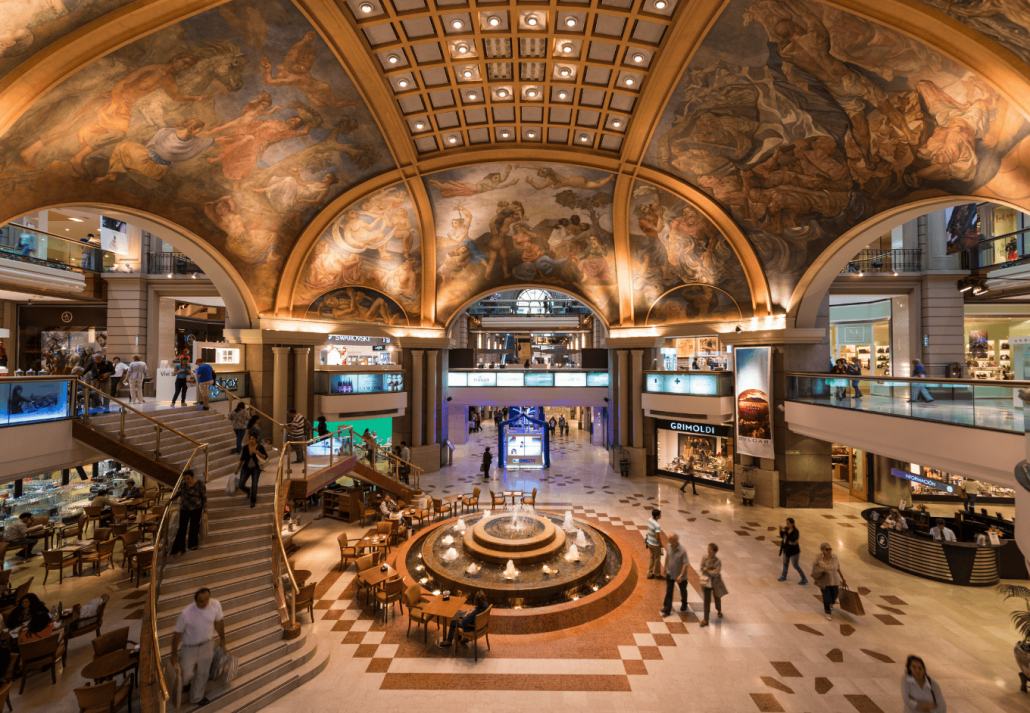 Shoppers wandering in an opulent mall with a grandiose ceiling mural.