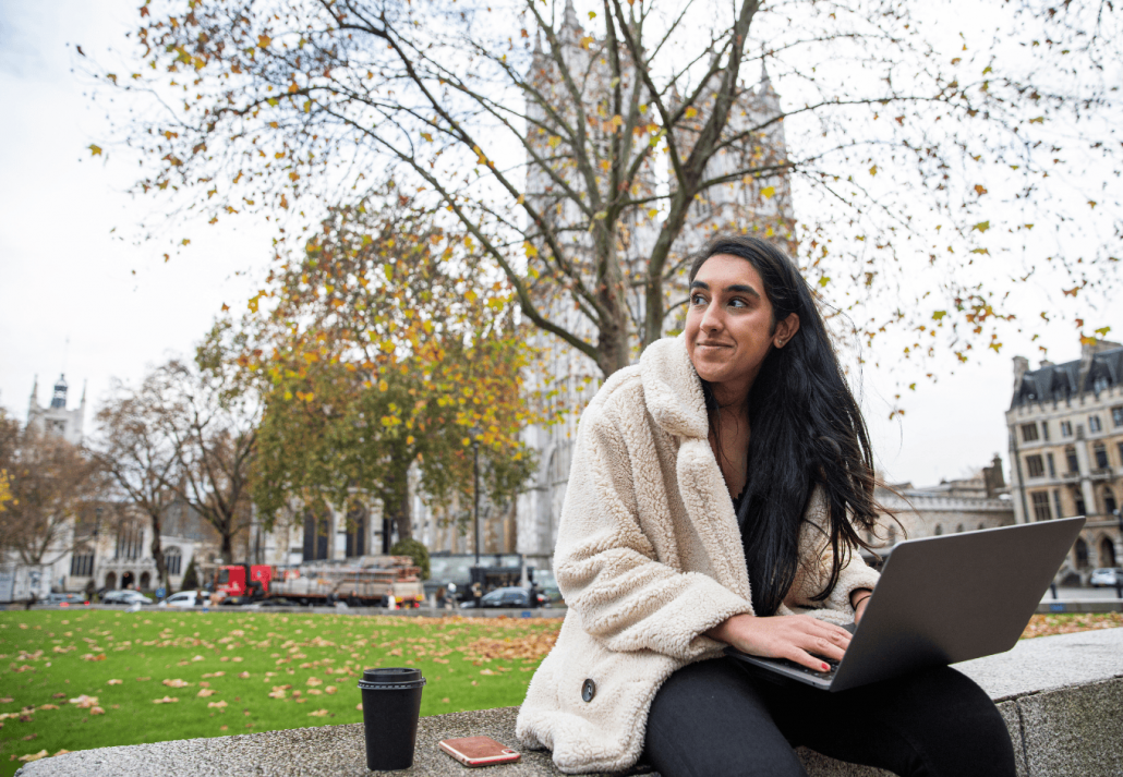 Indian girl works outdoors and uses her laptop, she drinks coffee and she is in a park.